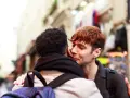 Un hombre joven con cabello rojizo susurra "bonjour" a su pareja, un momento íntimo en una calle parisina.