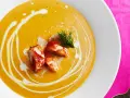 Un cremoso bisque de langosta, adornado con trozos de carne de langosta y un toque de eneldo, servido en un plato blanco sobre un mantel rosa vibrante.
