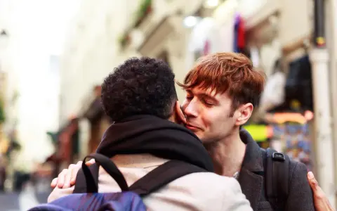 Un hombre joven con cabello rojizo susurra "bonjour" a su pareja, un momento íntimo en una calle parisina.