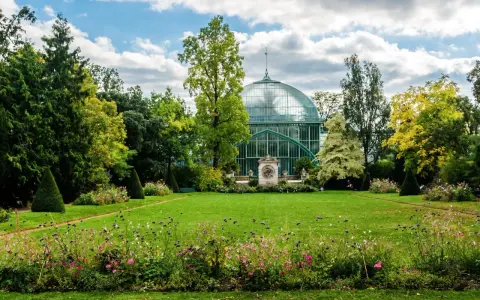 Un jardín exuberante en el Bois de Boulogne, con una gran estructura de cristal verde y un cielo azul con nubes.