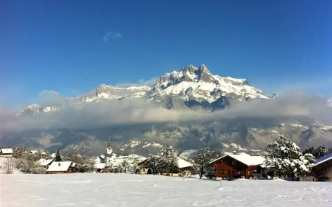 Un pueblo alpino cubierto de nieve, con casas de madera y una iglesia, bajo imponentes montañas nevadas. Un paisaje de invierno en francés.