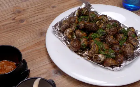 Caracoles con mantequilla y perejil, servidos en papel de aluminio sobre un plato blanco, junto a un cuenco con salsa.