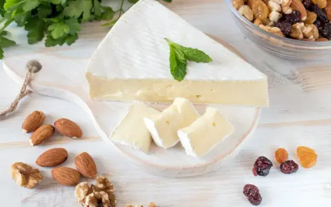 Queso brie en cuña y trozos, decorado con menta. Alrededor hay almendras, nueces y frutos secos. Ideal para quienes buscan información sobre queso brie calorias.