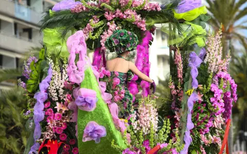 Una figura con un vestido floral y un tocado verde, rodeada de exuberantes flores rosas y moradas, participa en el **carnaval de Nice**.