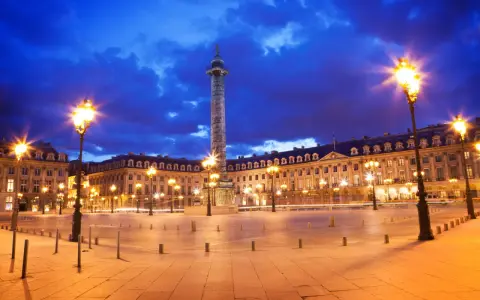 La majestuosa plaza Vendôme se ilumina bajo un cielo nocturno azul intenso, con farolas doradas y la imponente columna en el centro.