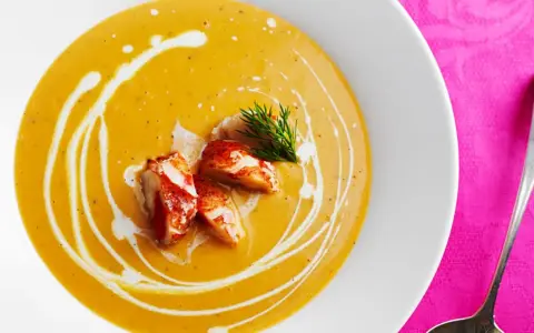 Un cremoso bisque de langosta, adornado con trozos de carne de langosta y un toque de eneldo, servido en un plato blanco sobre un mantel rosa vibrante.
