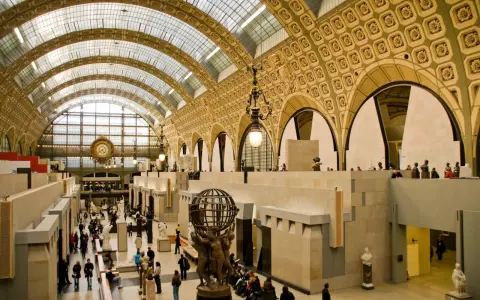 Interior del museo d'Orsay, con su impresionante techo de cristal y arcos dorados. Visitantes pasean entre esculturas y obras de arte.