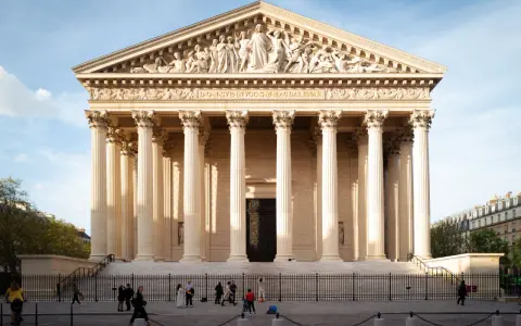 La imponente iglesia de la Madeleine en París, con sus columnas neoclásicas y esculturas en el frontón, se alza bajo un cielo azul.