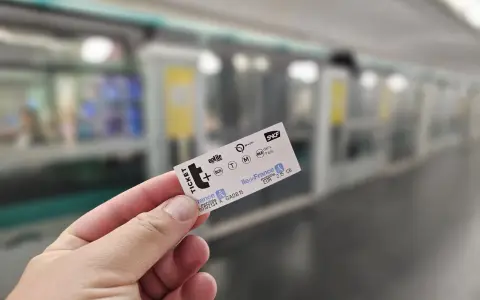Mano sostiene un ticket t+ para el transporte público en París, con un tren borroso al fondo.