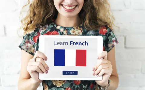 Una mujer sonriente sostiene una tablet que muestra "Learn French" y la bandera francesa. ¡Es hora de aprender como se habla en Francia!