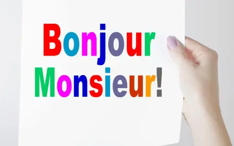 Una mano sostiene un cartel con "Bonjour Monsieur!" escrito en letras de colores.