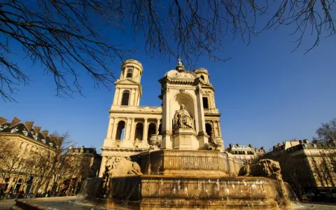 Fuente monumental frente a la iglesia de Saint Sulpice, con leones de piedra y una estatua central bajo un cielo azul claro.