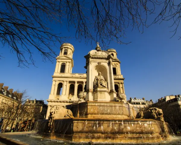 Fuente monumental frente a la iglesia de Saint Sulpice, con leones de piedra y una estatua central bajo un cielo azul claro.
