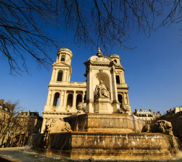 Fuente monumental frente a la iglesia de Saint Sulpice, con leones de piedra y una estatua central bajo un cielo azul claro.