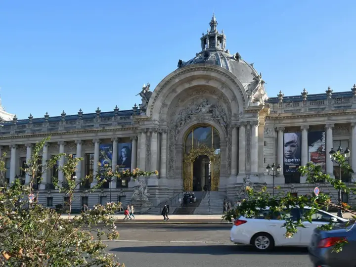 El Petit Palais, un edificio majestuoso con una cúpula y una entrada dorada, exhibe carteles de arte bajo un cielo azul.
