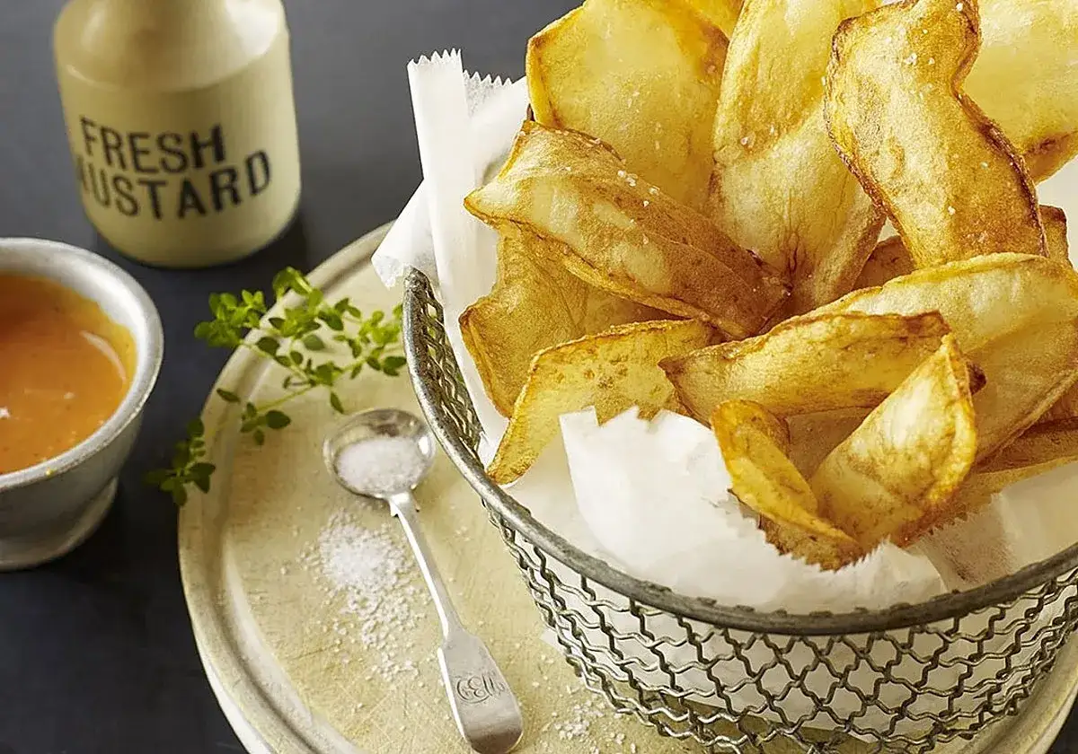 Crujientes patatas souffle recién hechas, servidas en una cesta metálica, acompañadas de salsa y sal marina.