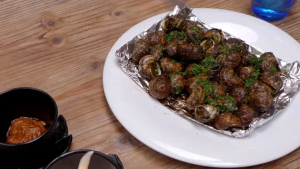 Caracoles con mantequilla y perejil, servidos en papel de aluminio sobre un plato blanco, junto a un cuenco con salsa.