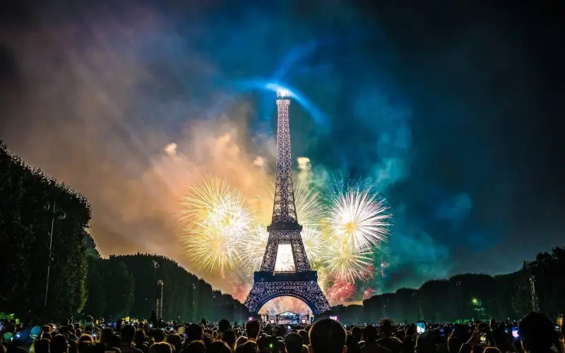 Espectacular espectáculo de fuegos artificiales ilumina la Torre Eiffel, celebrando porque se celebra el 14 de julio en Francia. Multitud observa.