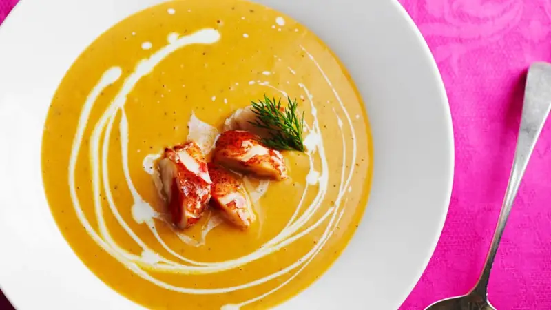 Un cremoso bisque de langosta, adornado con trozos de carne de langosta y un toque de eneldo, servido en un plato blanco sobre un mantel rosa vibrante.