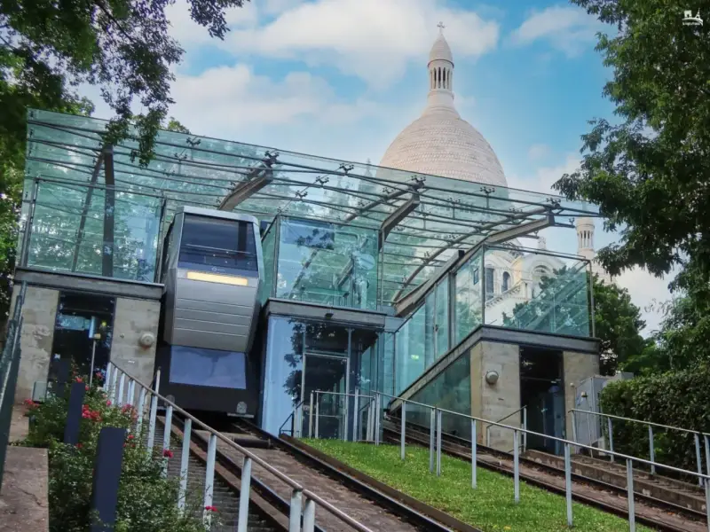 El funicular de Montmartre, con su moderna estación de cristal, asciende hacia la basílica del Sacré-Cœur bajo un cielo azul.