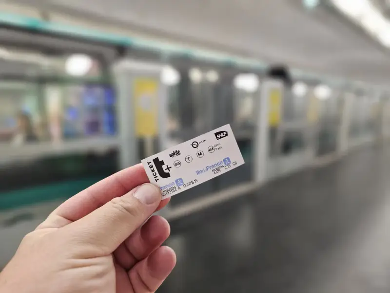 Mano sostiene un ticket t+ para el transporte público en París, con un tren borroso al fondo.