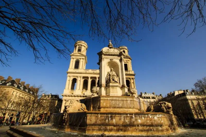 Fuente monumental frente a la iglesia de Saint Sulpice, con leones de piedra y una estatua central bajo un cielo azul claro.
